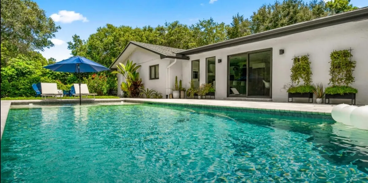 Turquoise swimming pool beside a white home with a shaded outdoor seating area